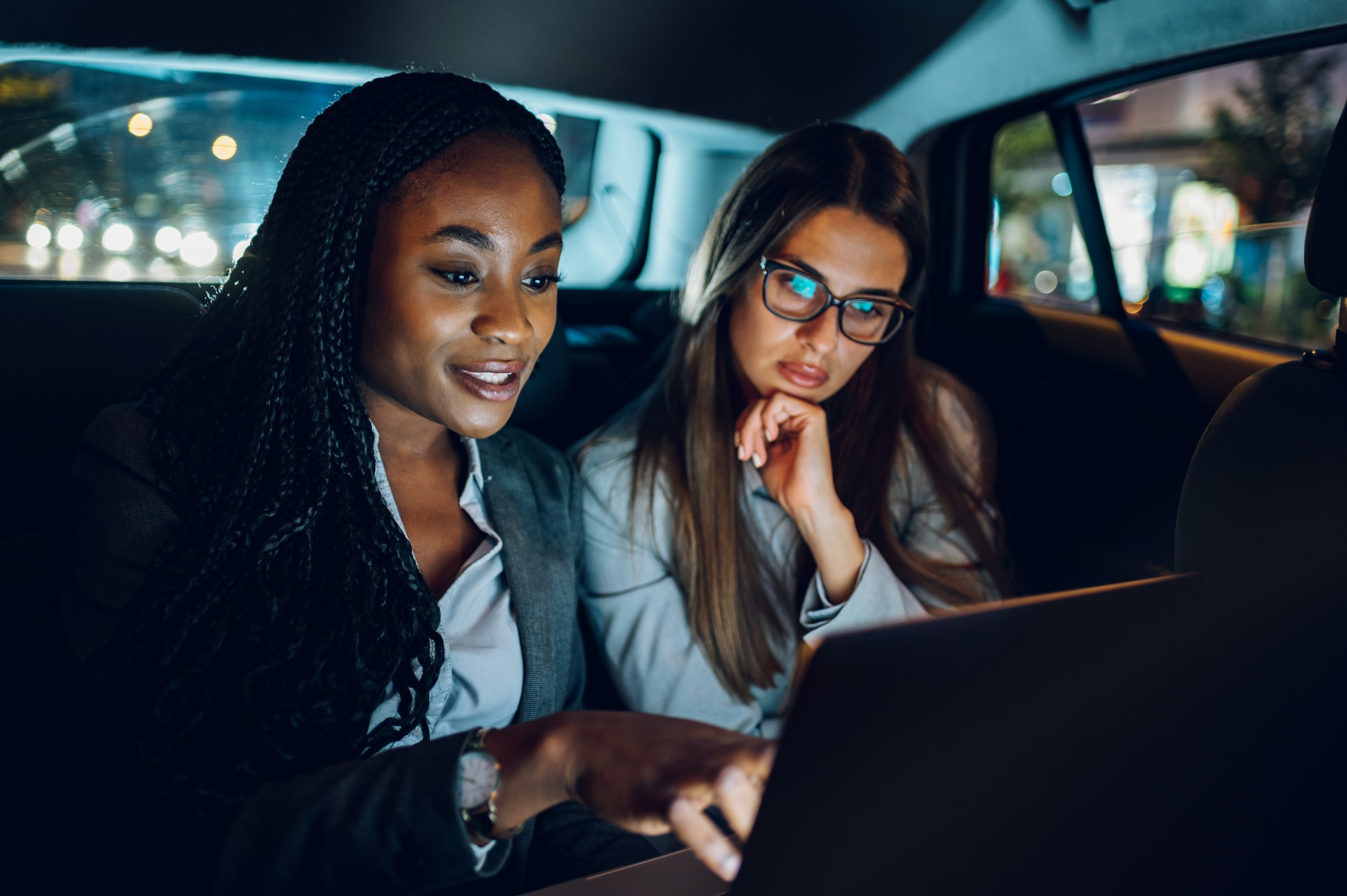Two multiracial business woman riding in a car and using a laptop